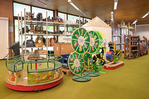 Innenaufnahme eines Museumsdepots mit bunten Karussellfiguren, zwei Glücksrädern, einem Kinderkarussell sowie Regalflächen mit Skulpturen, Archivkartons und historischen Geräten. Foto: Ingo Kröner.