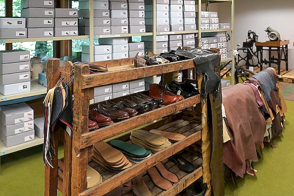 Innenaufnahme eines Lagerraums mit einem hölzernen Regal voller Musterschuhe, Lederproben und Einlegesohlen. Im Hintergrund stehen zahlreiche weiße Archivboxen in Regalen sowie eine alte Schuhmacher-Maschine. Foto: Ingo Kröner.